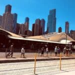 Queen Victoria Market's cream-roofed sheds under a clear blue sky, with Melbourne's city towers behind and people walking past a tram stop in the foreground