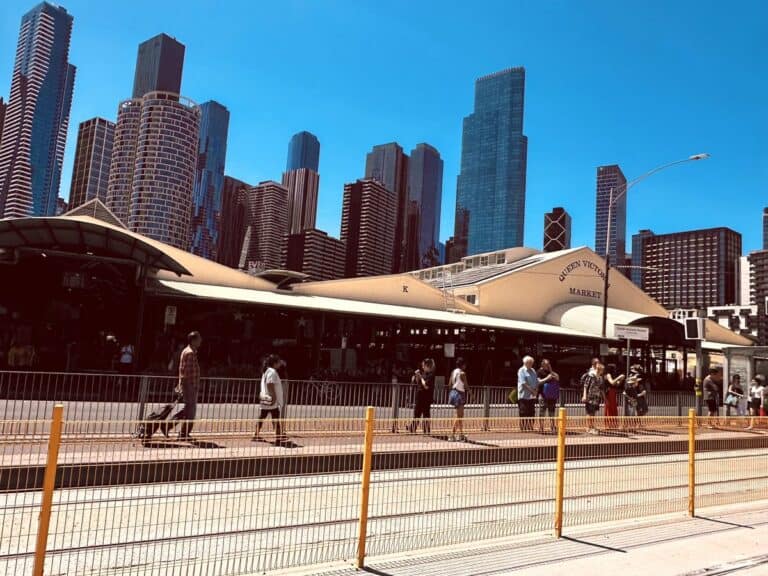Queen Victoria Market's cream-roofed sheds under a clear blue sky, with Melbourne's city towers behind and people walking past a tram stop in the foreground