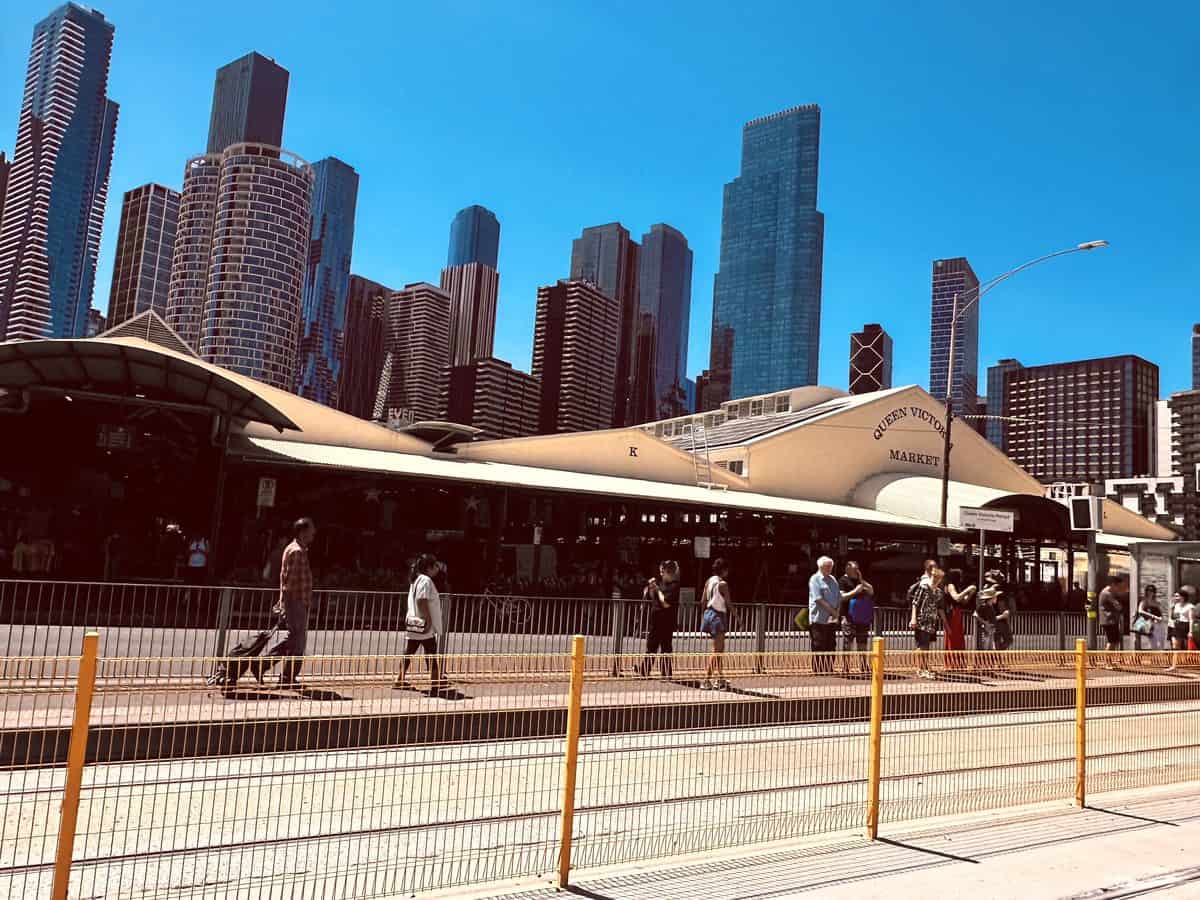 Queen Victoria Market's cream-roofed sheds under a clear blue sky, with Melbourne's city towers behind and people walking past a tram stop in the foreground